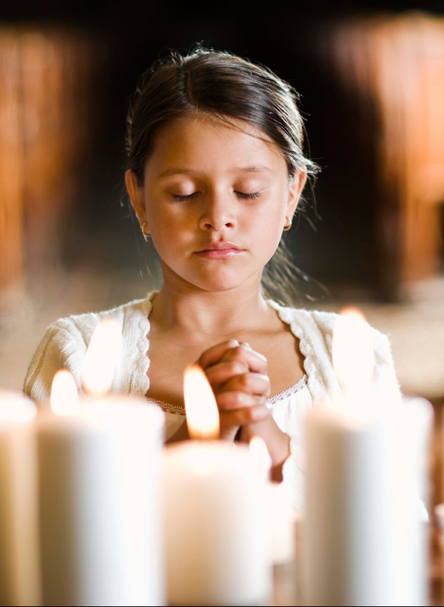 A brown-skinned young girl in a white dress and shawl kneels in front of several lit candles. Her eyes are closed and her hands folded in prayer.