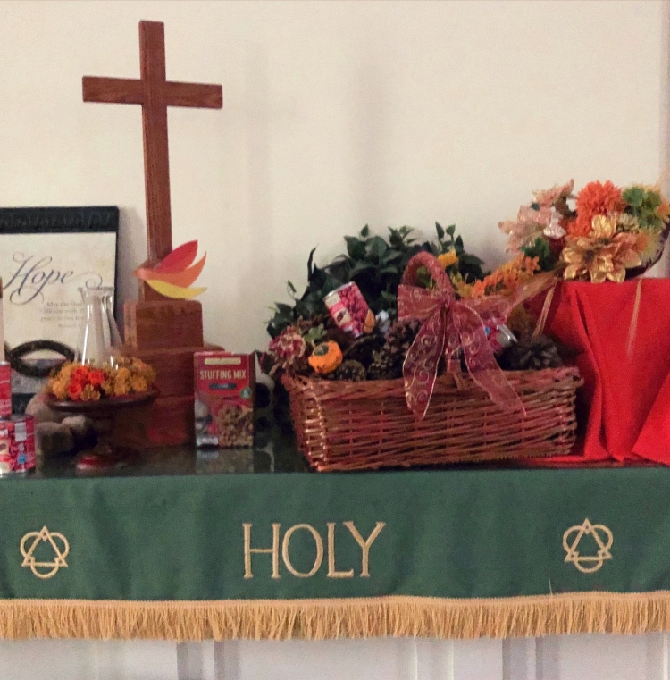 The East Bloomfield sanctuary altar is decorated with a harvest theme. On a green altar cloth reading "Holy" between geometric symbols of the Trinity are: a wooden altar cross, a piece of wall art including the word "Hope" and a Jesus fish, two floral arrangements, a flame-inspired dove, and food being collected for donations.