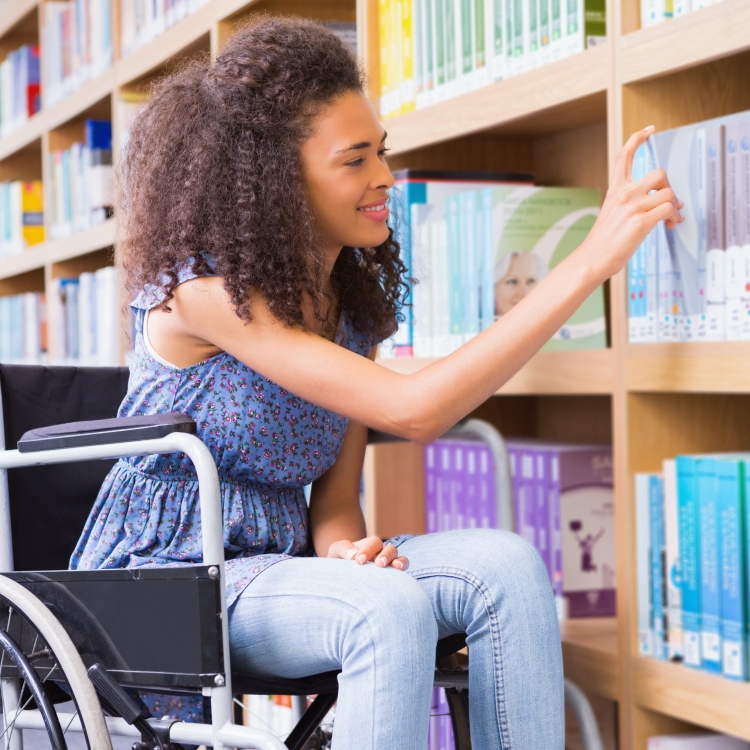 A light-skinned, young black woman with shoulder-length hair sits in a wheelchair. She is pulling a book off a library shelf in a well-lit room.